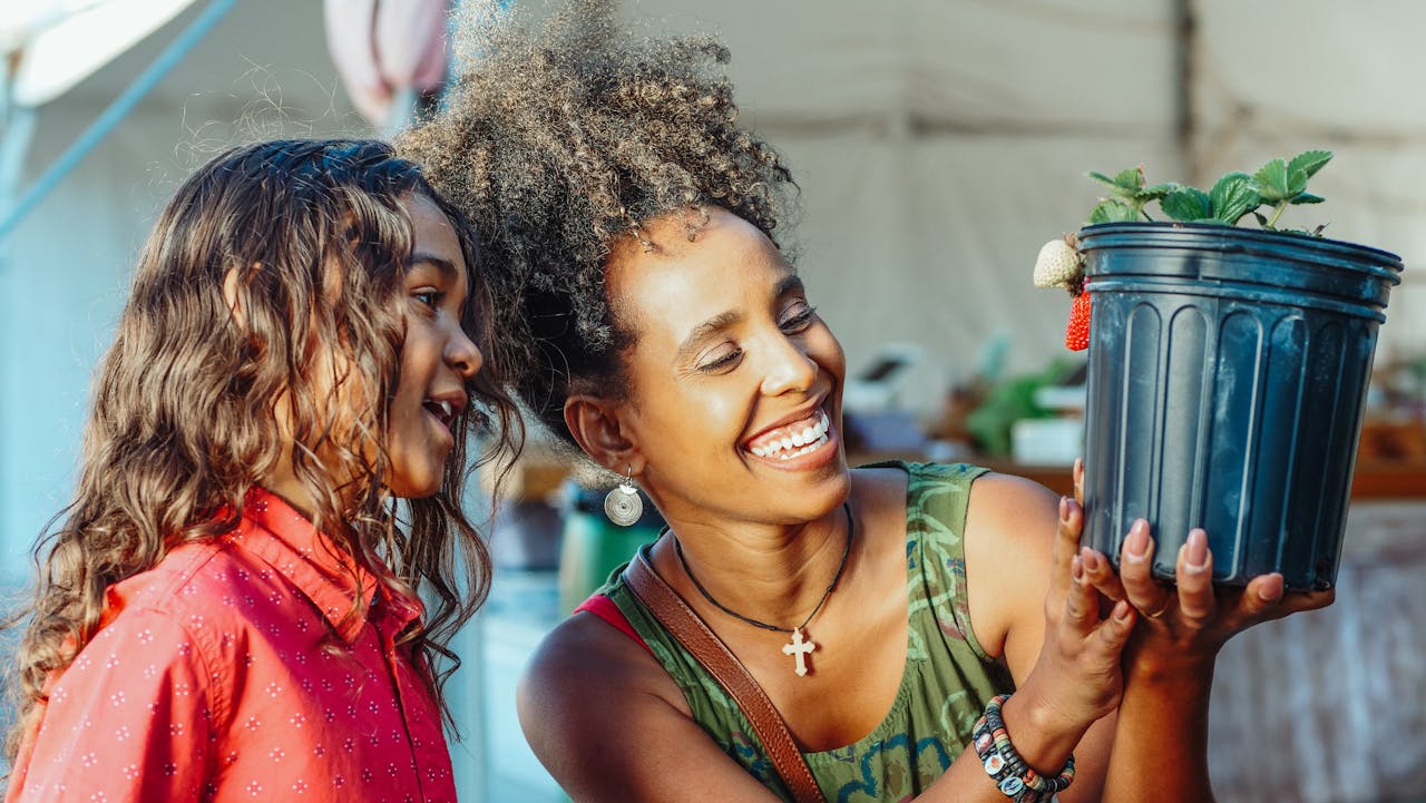 gallery-3 Mother and daughter smiling and holding a strawberry plant, showcasing happiness and togetherness.